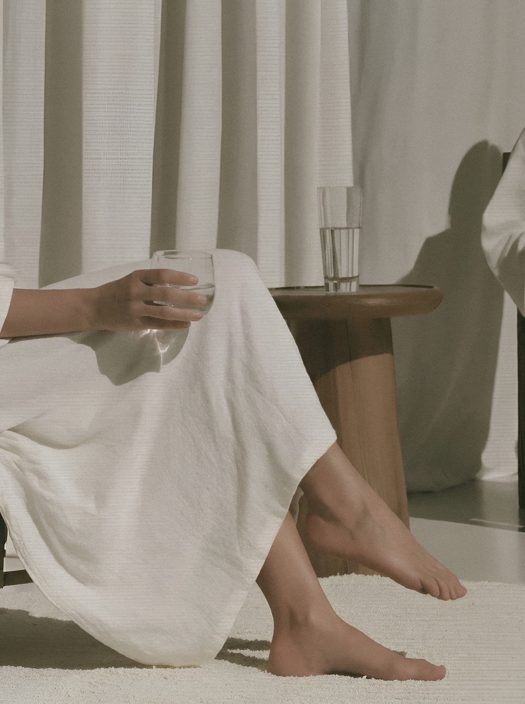 A hand holding a glass above white linen, with bare feet resting beside a sunlit curtain.