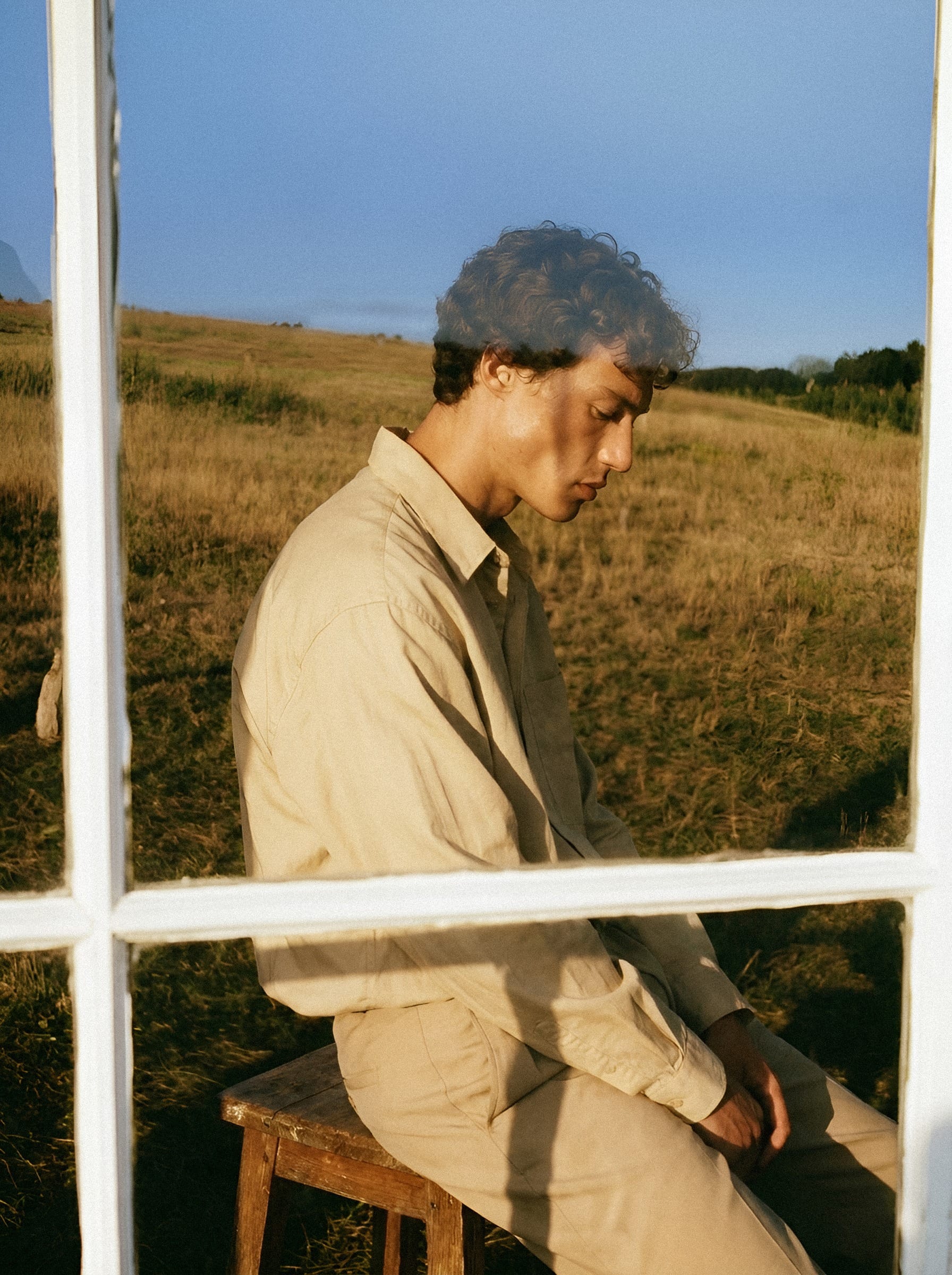 A man in a linen shirt seen beside an open window overlooking grassland.