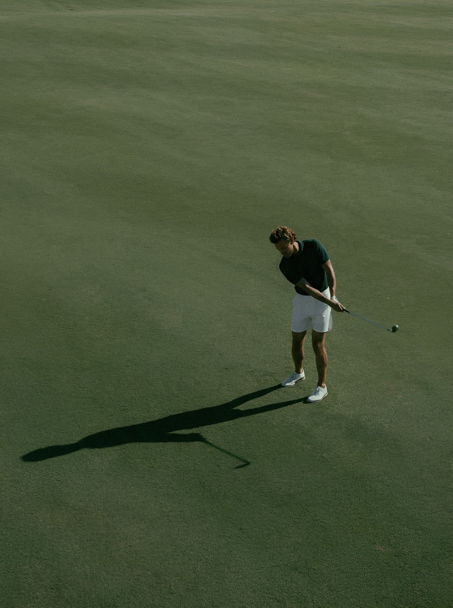A golfer standing over a ball, seen from above, with a long shadow across the green.
