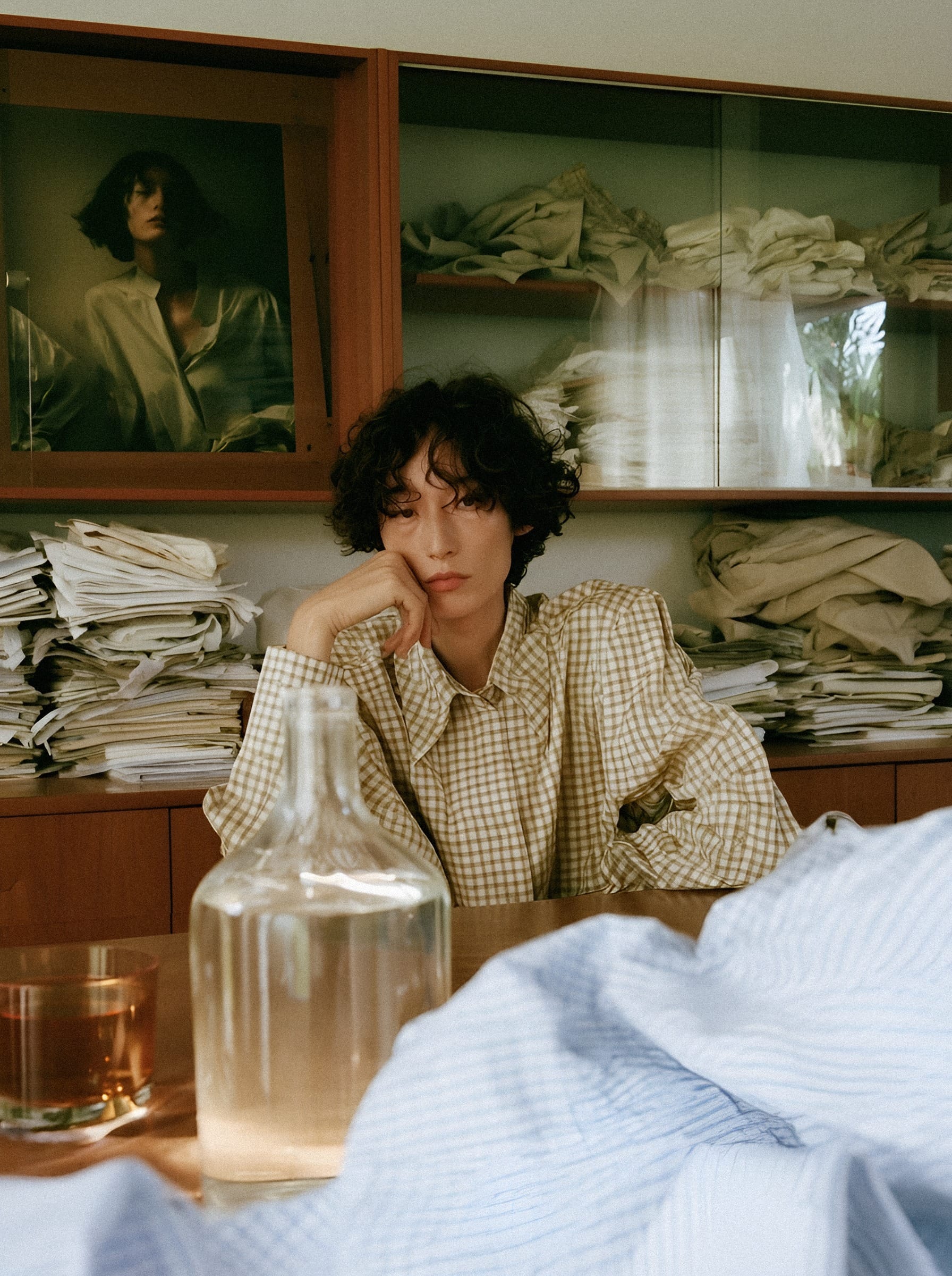 A woman in a checked shirt seated before shelves of folded linen.