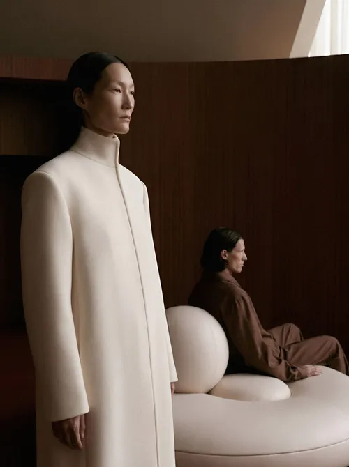A woman in a cream coat beside curved pale seating and a seated figure against wooden walls.